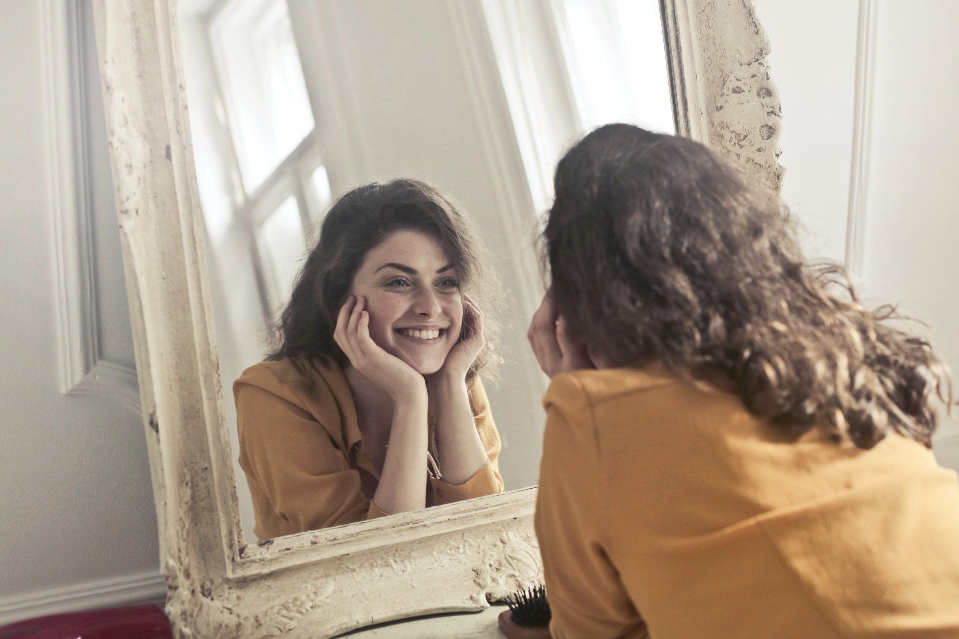 Woman smiling at her reflection in an ornate mirror, resting her chin on her hands.